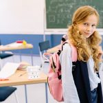 A young girl with a backpack stands confidently in a bright classroom setting.