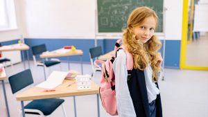 A young girl with a backpack stands confidently in a bright classroom setting.